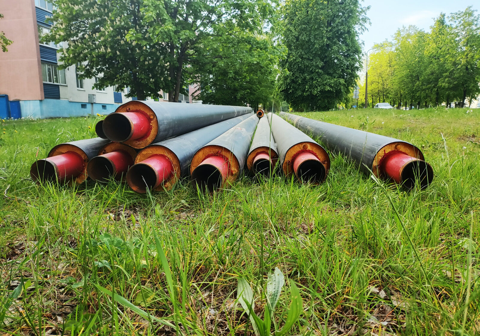 Large diameter insulated pipes, black exterior, red interior, thick orange thermal insulation, placed horizontally on vibrant green lawn. Apartment building and lush green trees form backdrop on sunny day.